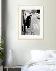 A framed wall art black-and-white photograph of an elderly person with a cane stepping onto a street hangs above a white bed with pillows; a plant and white vase sit on a small table nearby, creating mindful home decor.