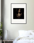 A fine art print of a person’s face with dark hair hangs above a bed with white bedding, creating a conscious interior design. A small table with a white vase and green plant sits beside the bed.