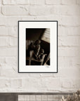 A fine art print in black and white, featuring a person sitting with knees drawn up, is displayed as framed wall art above a light-colored mantle adorned with a small plant and decorative objects—perfect for conscious interior design.