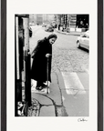 An elderly woman with a cane crosses a cobblestone street in this black and white fine art print. She wears a long coat and headscarf, with cars and buildings in the backgroundβa thoughtful piece for conscious interior design.