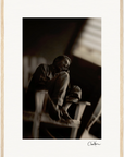 A sepia-toned fine art print of a man sitting on a wooden chair with knees drawn up, looking slightly upward. Light streams in from a window above, illuminating the dark background. Finished with a light wooden border, it’s perfect for mindful home decor.