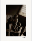 A man sits barefoot on a wooden chair in a dimly lit room, resting his cheek on his hand. This black and white fine art print is signed “Chan Xaru” at the bottom right corner, perfect as framed wall art for sophisticated interiors.