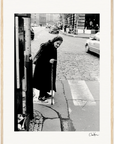 An elderly woman wearing a headscarf and dark clothing uses a cane to cross a cobblestone street. Cars are in the background, and pedestrians walk on the sidewalk. This black-and-white scene adds mindful home decor appeal to your space.