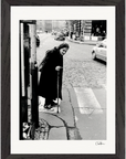 An elderly woman with a cane stands at a crosswalk on a cobblestone city street, captured in striking black and white. Perfect as inspirational wall art, this photo comes with a gallery-quality frame and a dark border for conscious interior design.