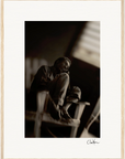 A man sits barefoot on a wooden chair with one knee raised, gazing thoughtfully to the side in a dimly lit room. This fine art print is framed with a white mat and light wood frame, featuring a signature at the bottom—perfect framed wall art.