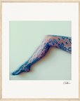 A single outstretched leg in black lace-patterned tights with a floral design is photographed against a light background. The image features a gallery-quality frame and has a signature in the bottom right corner.