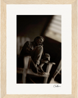 A black-and-white photo shows an older man sitting on a wooden chair with his legs up, head resting on his hand. The blurred background and dim lighting enhance this fine art print, making it perfect for mindful home decor. Displayed in a light wood frame.