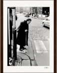 An elderly woman with a cane crosses a cobblestone street at a crosswalk in an urban setting, captured as cars and pedestrians move in the background—a perfect fine art print for conscious interior design.