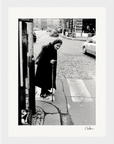 An elderly woman in dark clothing leans on a cane while crossing a cobblestone street, cars and buildings lining the background. This black and white fine art print is perfect for adding mindful home decor or framed wall art to any space.