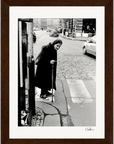 An elderly woman in a headscarf and dark clothing leans on a cane at a crosswalk on a cobblestone city street. Cars and buildings form the backdrop, making this scene perfect as inspirational wall art for mindful home decor or display in a gallery-quality frame.