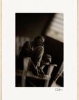 A man sits barefoot on a wooden chair indoors with one knee raised, gazing upward. The black-and-white photo features soft focus, dramatic lighting, and a visible signature—ideal as framed wall art or mindful home decor.