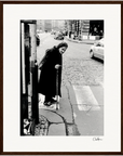 An elderly woman with a cane walks slowly across a cobblestone street at a crosswalk, framed by cars and old buildings in the background. This black and white image makes striking framed wall art for mindful home decor.