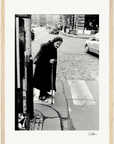 An elderly woman with a cane walks slowly across a cobblestone street at a crosswalk. She wears a long coat and headscarf. Cars and people move in the background, creating an evocative scene ideal for a fine art print or mindful home decor.