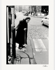 An elderly woman with a cane stands at a street corner, looking down as she prepares to cross a cobblestone road. Cars line the street and historic buildings rise behind her. This black and white scene makes an evocative fine art print or inspirational wall art.