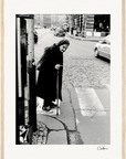 An elderly woman with a headscarf and cane stands at the edge of a cobblestone street, ready to cross. Cars and people appear in the background. This black-and-white image is framed in light wood, perfect as inspirational wall art for conscious interior design.