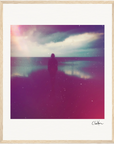 A silhouetted figure stands on a beach at sunset under a cloudy sky. The dreamy image, with pink and purple tones and a soft, grainy effect, is framed in light wood—perfect for mindful home decor.