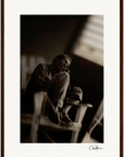 A sepia-toned fine art print shows a man sitting barefoot on a wooden chair with his knees pulled up, gazing to the side. Light from a window softly illuminates the dark background—a mindful home decor piece signed at the bottom right.