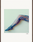 A single leg in blue lace floral-patterned stockings extends diagonally on a light background. This framed wall art features a thin brown border and is signed Clarkyan, making it a beautiful addition to mindful home decor.