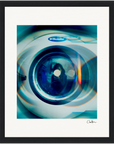 A close-up photo of an Electrolux washing machine door with blue-toned light reflections and circular lens effects, presented as inspirational wall art in a gallery-quality frame with a white mat and artist’s signature at the bottom right.