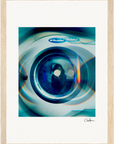 A close-up view of the front window of a washing machine with a blue and green color filter and circular light reflections. The image is presented as framed wall art with a gallery-quality frame and features a small signature at the bottom right.