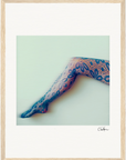 A single outstretched leg in patterned floral lace tights is set against a pale background with a soft, blurred effect. Framed in a gallery-quality frame, this piece brings mindful home decor to any conscious interior design. An artist’s signature appears below.
