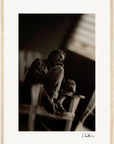 A black and white photo of a man sitting on a chair indoors, knees drawn to his chest, looking off to the side. The dimly lit background and gallery-quality frame make it perfect for mindful home decor; signed at the bottom right.