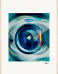 A close-up of the front door of an Electrolux washing machine, with blue and orange light reflections, framed in white with a thin beige border. The artist's signature appears in the bottom right corner, perfect as mindful home decor or a fine art print.