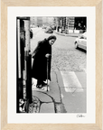 An elderly woman in a long coat and headscarf walks with a cane at a crosswalk on a cobblestone street. This black and white fine art print is elegantly framed with light wood, perfect for conscious interior design.