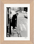 A black and white photo shows an elderly woman with a cane at the edge of a city street near a crosswalk, wearing a headscarf and long coat. Cars and buildings form the background, making this image ideal as a fine art print for conscious interior design.