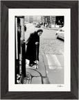 Black and white fine art print of an older woman with a cane at a crosswalk on a cobblestone street, cars and buildings behind her. Presented as inspirational wall art in a dark wooden frame.