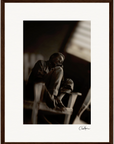 A sepia-toned fine art print captures an older man sitting barefoot on a wooden chair, one knee drawn up, gazing thoughtfully into the distance. This inspirational wall art is elegantly framed with a white mat and dark wood.