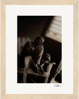 A black-and-white photo of a person sitting on a chair with knees pulled to their chest, looking to the side; this fine art print is displayed in a gallery-quality light wood frame and signed at the bottom right.