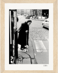 Black and white photo of an elderly woman with a cane crossing a cobblestone street; cars and buildings in the background. Her long coat and headscarf evoke timeless strength, perfect as inspirational wall art or mindful home decor.