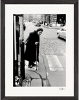 An elderly woman in a dark coat and headscarf walks with a cane along a cobblestone street, pausing at a crosswalk. Cars and buildings fill the background. This black-and-white photo is presented as refined framed wall art.
