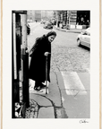 An elderly woman in a headscarf crosses a cobblestone street with her cane at a crosswalk. Cars and buildings line the background. This black and white fine art print adds timeless character to mindful home decor or inspirational wall art displays.