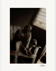 A man sits on a wooden chair with his knees drawn up, looking off to the side. The black and white photo, featuring soft, blurred edges and dramatic lighting, comes in a gallery-quality frame—perfect for conscious interior design. A signature appears bottom right.