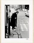 An elderly woman in a headscarf and coat uses a cane to cross a cobblestone street, with cars and pedestrians nearby. This scene captures timeless resilience, perfect as an inspirational wall art or fine art print in a gallery-quality frame.