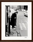 A black and white photo shows an elderly woman with a cane standing at a street corner in a long coat and scarf. Cars and buildings line the cobblestone road, creating stunning framed wall art perfect for mindful home decor.