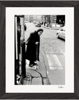 An elderly person in a dark coat and headscarf uses a cane while stepping onto a cobblestone street at a crosswalk, with cars in the backgroundβa striking image perfect for framed wall art that enhances mindful home decor.