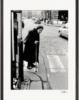An elderly woman with a cane stands at the edge of a cobblestone street, preparing to cross. Cars and pedestrians are visible in the background. This black and white photo, set in a gallery-quality frame, enhances any conscious interior design.