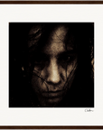 A grainy, high-contrast portrait of a person with long, dark hair partially covering their face, looking directly at the camera. Framed with a dark border and signed in the corner, this piece brings mindful home decor to conscious interior design.