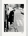 An elderly woman with a cane pauses at a cobblestone crosswalk, city cars passing by. Wearing a long coat and scarf, she is captured in black and white. This signed fine art print adds mindful home decor to any space.