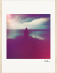 A silhouetted person stands by a body of water under a cloudy sky, with soft pink and purple light effects and a grainy texture. The fine art print features a gallery-quality frame, light wooden border, and signature in the bottom right corner.