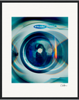 A close-up photo of the front door of an Electrolux washing machine, featuring layered blue and teal light effects. Presented as a fine art print in a gallery-quality frame, the image is signed in the bottom right corner.
