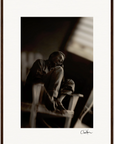 A black and white photo of an older man sitting sideways on a wooden chair with his knees drawn up, gazing off to the side. The dark, blurred background enhances the mood. An ideal fine art print, complete with a visible signature at the bottom right.