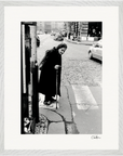 An elderly woman with a cane stands at the edge of a cobblestone street, ready to cross. She wears a dark coat and headscarf. Cars and buildings line the background. This black-and-white fine art print is presented in a gallery-quality frame.
