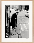 An elderly woman with a cane crosses a cobblestone city street at a crosswalk. She wears a long dark coat and headscarf. Cars and buildings are visible in the background. This black and white photo makes striking framed wall art.