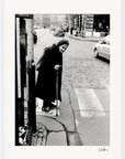 An elderly woman with a cane crosses a cobblestone city street at a crosswalk, wearing a dark coat and headscarf. This fine art print adds character and thoughtful beauty to any space, perfect for mindful home decor.