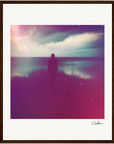 A silhouetted person stands near a calm body of water under a cloudy sky, with pink and purple light effects and a grainy texture. The image is presented as a fine art print in a simple brown gallery-quality frame, signed at the bottom right.