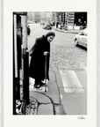 An elderly woman in a headscarf and dark coat uses a cane to cross a city street, with cars in the background. This black and white image makes striking framed wall art, perfect for mindful home decor or gallery-quality framing.